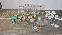 Wide view of various clear glass jars with mixed metal and plastic lids arranged on table with the small white metal rack and cleaning cloths.