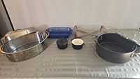 Wide shot of the seven cookware and bakeware items on a table; stainless steel roasting pan with lid, blue ceramic baking dish, Pyrex casserole with wooden holder, heavy duty black pot with lid, two small bakeware items (one with lid).