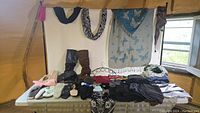 Wide view of table with clothing and footwear items displayed including scarves hanging above, boots, slippers, socks, leggings, and t-shirts on the table.