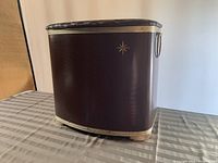 Front left angle view of mid century brown laundry hamper with gold star logo and handle, sitting on table.