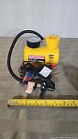 Yellow portable inflator with pressure gauge and hose, car lighter plug shown with ruler for size.