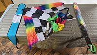 Checkered black and white Delta-shaped kite with rainbow edges and a blue carry case, displayed unfolded on a surface next to a packaged colorful kite.