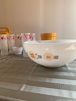 Side view of large Pyrex mixing bowl with orange-brown floral pattern, Tom Collins glasses, yellow Tupperware ice bucket and yellow accessory in background.