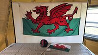 Setup with a large 5' x 3' Welsh flag hanging on wall behind a table with a smaller 18" x 12" Welsh flag, Welsh cap, and small magnet displayed on the table.