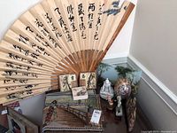 Large Asian folding fan displayed upright with visible calligraphy and painting, surrounded by various decorative objects including a painted tray, figurines, and coasters.