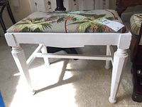 Front view of the painted bench with tropical leaf and pinecone pattern upholstery showing the entire bench structure and some scratches on the legs and frame.