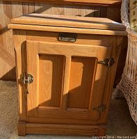Wooden cabinet with a single door with recessed panels, metal hinges, and latch. Natural wood finish with visible scratches. Shows side and front view with sunlight casting shadows.