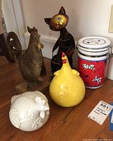 Photo showing the metal hen, yellow ceramic hen, white ceramic hen, metal cat sculpture, and red and white canister grouped on a wooden surface.