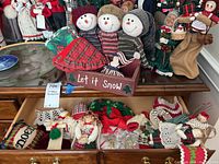 Wooden box with three snowmen inside and assorted Christmas ornaments displayed on a table with other holiday decorations in background.