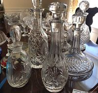 Four clear glass decanters of varying shapes with intricate cut patterns, placed on a wooden surface with other glassware visible in background.