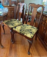 Two wooden dining chairs with fabric upholstered seats and carved backs shown positioned side by side on hardwood floor.