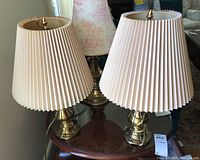 Two brass table lamps placed on a wooden table, showing pleated beige fabric lampshades and brass bases.