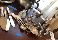 Wide view of the silver spreader knives with mother-of-pearl handles, perfume bottle, and silver goblet alongside a silver pitcher showing ornate handle