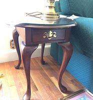 Side view of dark wood side table showing glass top, drawer with brass handle, and cabriole legs near a green sofa.