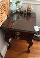 Side table showing glass top, two drawers with brass handles, curved legs, and polished dark wood finish.