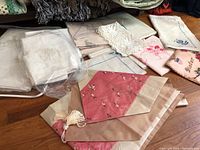 A selection of table linens including white lace and embroidered cloths along with colored pieces with tassels and floral embroidery.