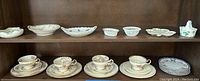 Four tea cups with matching saucers and various decorative bowls and plates on a brown wooden shelf.