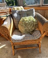 Wicker chair with geometric pattern cushions and green ruffled decorative pillow shown in indirect sunlight, visible some unraveling on armrest.