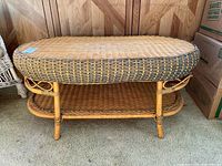 Full view of oval wicker coffee table showing top surface, legs, and lower shelf. Some unraveling visible on top edge.