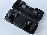 BOSE wireless earbuds seated in open charging case. Case interior shows left and right indicators and charging connectors. Earbuds are black with ear tips and winged tips for secure fit.