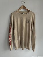 Beige Kanye West Kid Cudi sweatshirt hanging on hanger showing front side with black and red text.