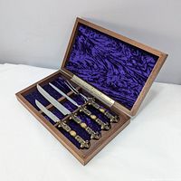 Open walnut box lined with purple velvet showing four pieces of carving utensils with ornate handles.