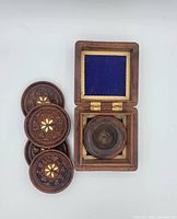 Photo showing four carved wooden coasters with floral inlay placed next to a rectangular wooden box with open hinged lid and blue felt lining inside.