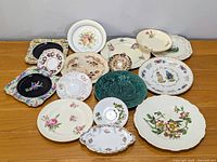 Top-down photo showing the complete assortment of 16 plates and dishes arranged on a table. Various sizes with floral and bird motifs in cream, white, green, and black color tones.