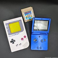 Overview of original Game Boy DMG-01, blue Game Boy Advance SP and Super Mario Land cartridge on black background