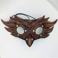 Front view of brown leather owl mask showing reflective lenses and stylized wing feather shapes extending from sides.
