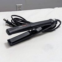 Black hair straightener with attached power cord on white sheet background.