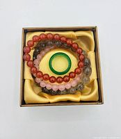 Top-down view of set in presentation box showing jade ring placed inside three stacked bracelets of carnelian, rose quartz and rutilated quartz beads on yellow satin liner.