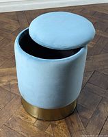 Straight view of modern velvet blue ottoman stool with lid lifted to show hollow storage interior and shiny gold base on wood floor.