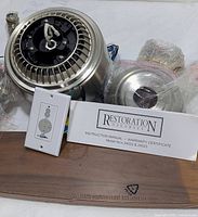 Close-up photo showing brushed metal housing of fan motor, white wiring, remote control with wall mount plate, instruction manual and warranty certificate labeled Restoration Hardware Model AC-552(24022). Wooden fan blade beneath.