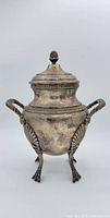 Front view of antique silver urn showing lid with pinecone finial, curved handles, and claw feet with shell motifs.