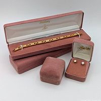 Open long pink velvet box containing gold-tone link bracelet with clear stones, two closed long boxes underneath, open small box with stud earrings, closed small box