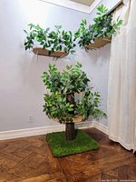 Overall view of the assembled cat tree with the two wall-mounted shelves in the corner of a room, showing artificial green leafy branches around the platforms and turf base.