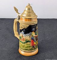 Front diagonal view of the beer stein showing a man and woman in Bavarian dress with scenic mountain and village background. Lid is closed.