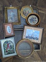 Multiple small framed artworks displayed inside vintage suitcase including Victorian era portraits, floral embroidery, stained glass style art, and maritime scenes.