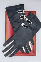 Full view of both black leather gloves laid on red box to show overall condition and decorative white bows on the cuffs.