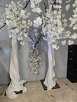 Photo showing three tall artificial cherry blossom trees with white flowers, fabric-wrapped trunks, and square black metal bases against a concrete wall.