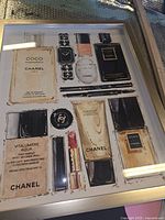 Framed art print showing an artistic display of Chanel brand products including perfume bottles, lipstick, makeup, and a watch.