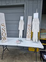 Photo of two white wooden nutcracker decorations and two wooden SNOW signs on a white folding table against a gray roll-up door.