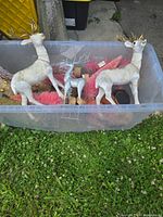 Two large light-colored reindeer figurines with gold antlers, a small silver glitter reindeer, pink feathery sprays, and other assorted decorations in a plastic bin outside on grass