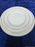 Stack of three plates of different sizes, white with gold trimming around the rims on a blue cloth