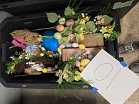 Black plastic bin box filled with various Easter decorations including fabric bunnies, floral artificial arrangements, wooden planters, and pastel-colored eggs on garlands.