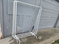 White heavy duty metal clothing rack photographed outside against weathered wooden wall and grey garage door, showing stable rectangular frame with lower cross bars and wide feet.