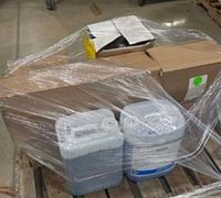 Two large plastic gallon containers shrink-wrapped on a wooden pallet along with cardboard boxes behind them.