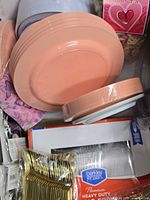 Stack of peach color plates with some clear and gold disposable forks in packaging inside bins