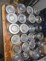 Top view showing multiple unopened metal cans with pull-tab lids, arranged in a cardboard box on a wooden surface.
